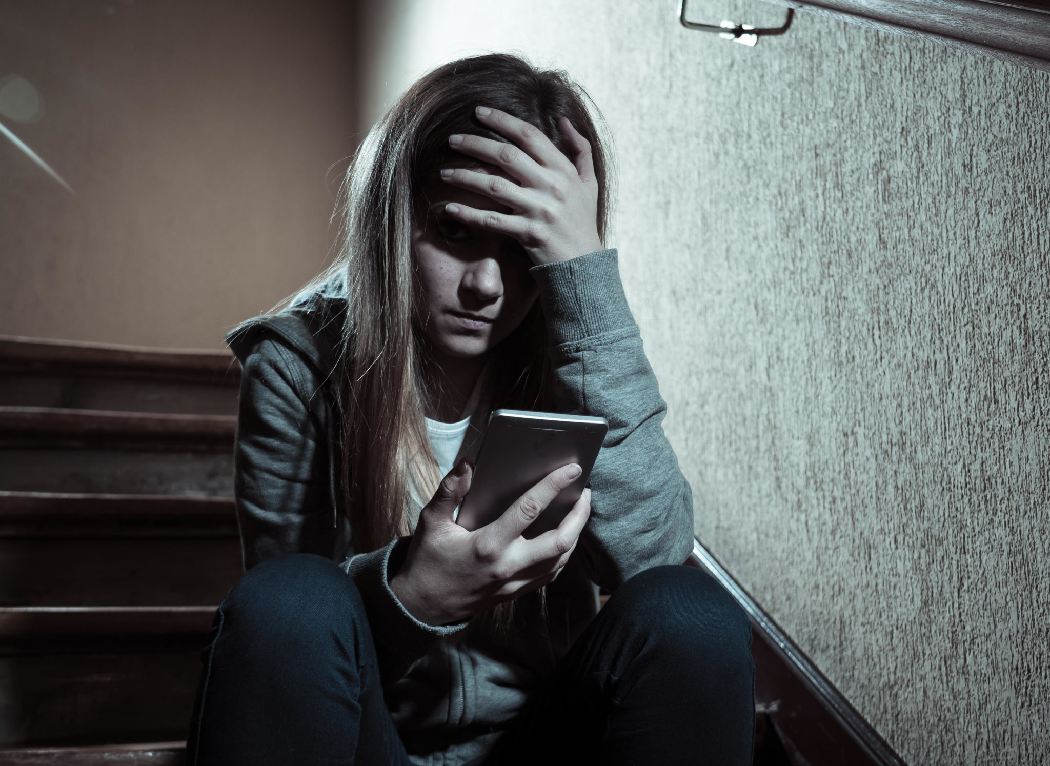 Teenager girl victim of online stalker suffering from cyberbullying abuse feeling lonely and hopeless sitting on stairs with dark light, 20-03-2019. Image: Samuel wordley / Alamy