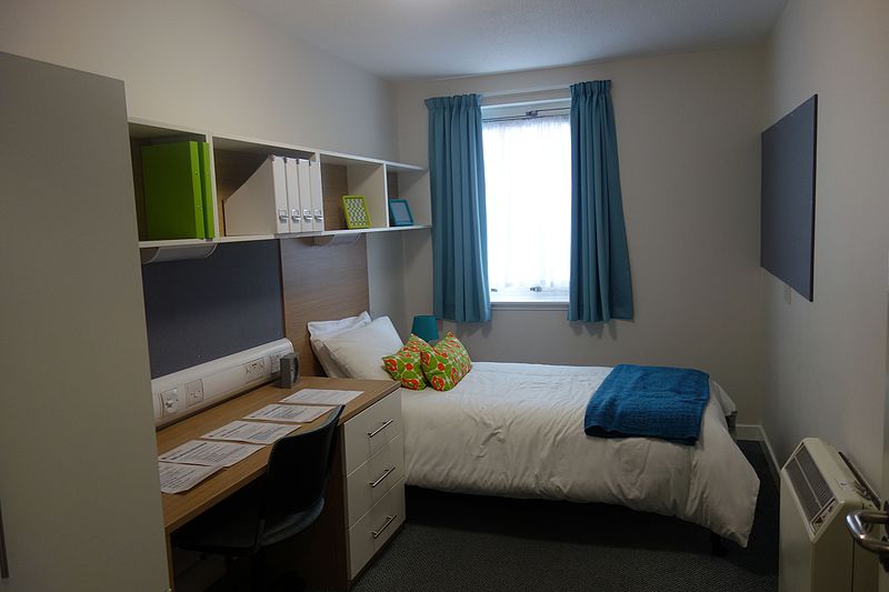 digs. Image shows a small room with a desk, overhead shelves, a bed and a window with curtains open.