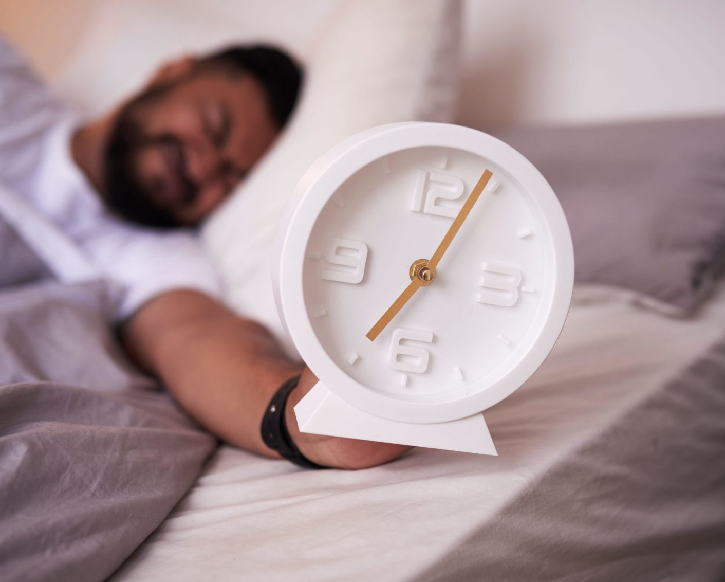 A 5am clock face in focus with an exhausted man sleeping in bed.