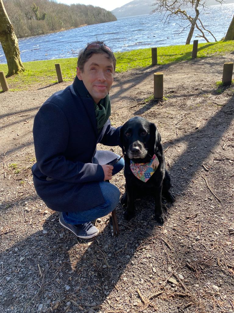Martin Gordon crouching on the ground beside his guide dog in the park.