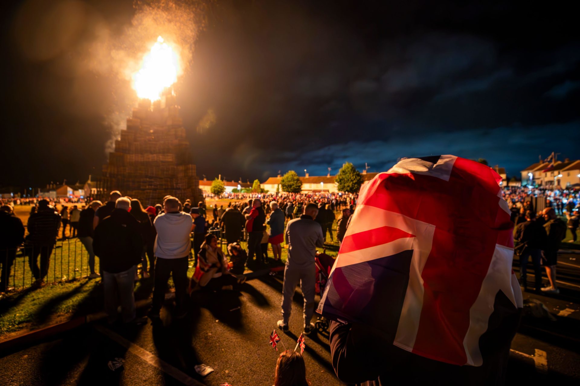 People watch the burning of the loyalist Corcrain bonfire in Portadown, Co Armagh. One person carries a British flag on their back.