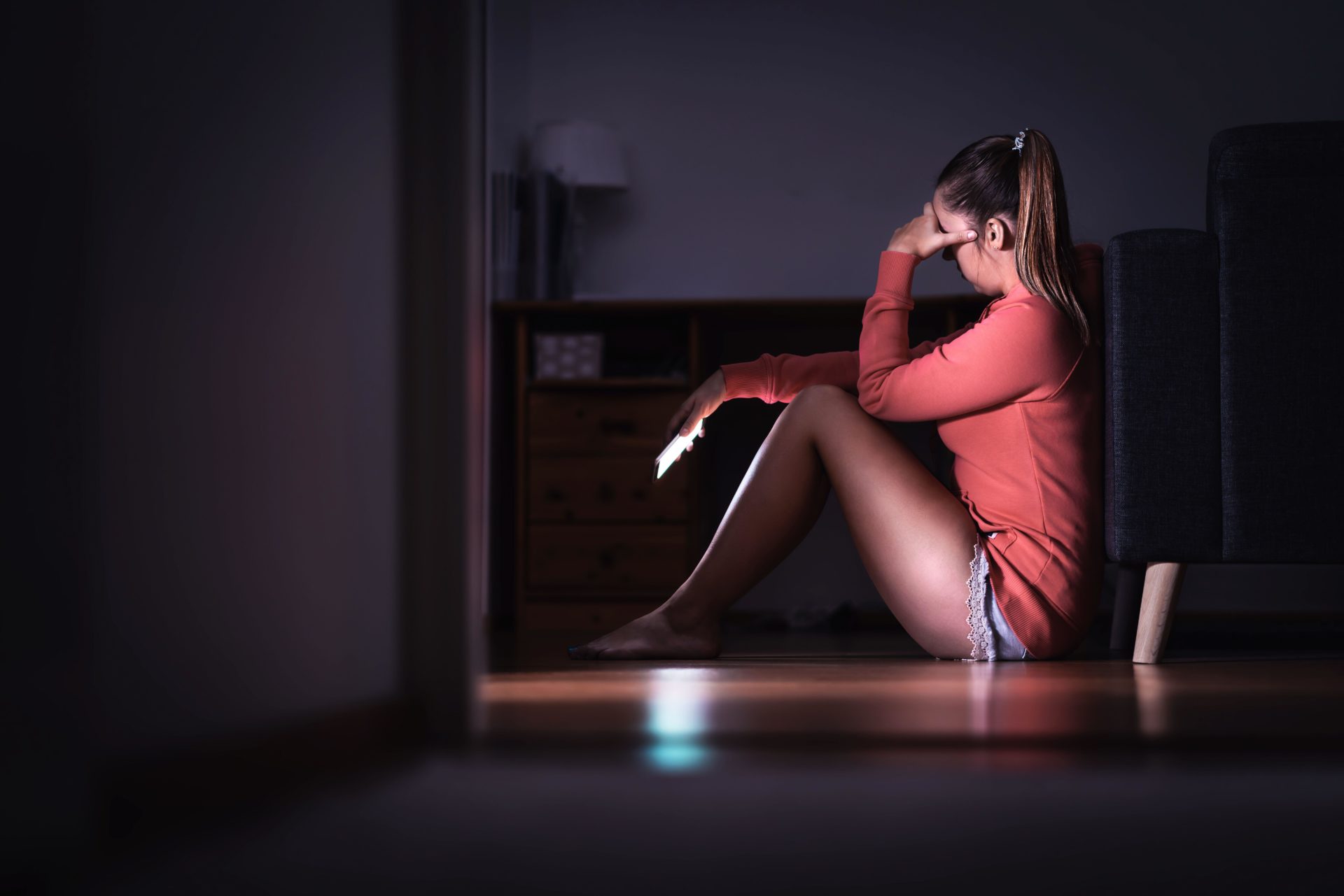 A woman holding her head with her hand, sitting on the floor in her pyjamas while holding a phone.