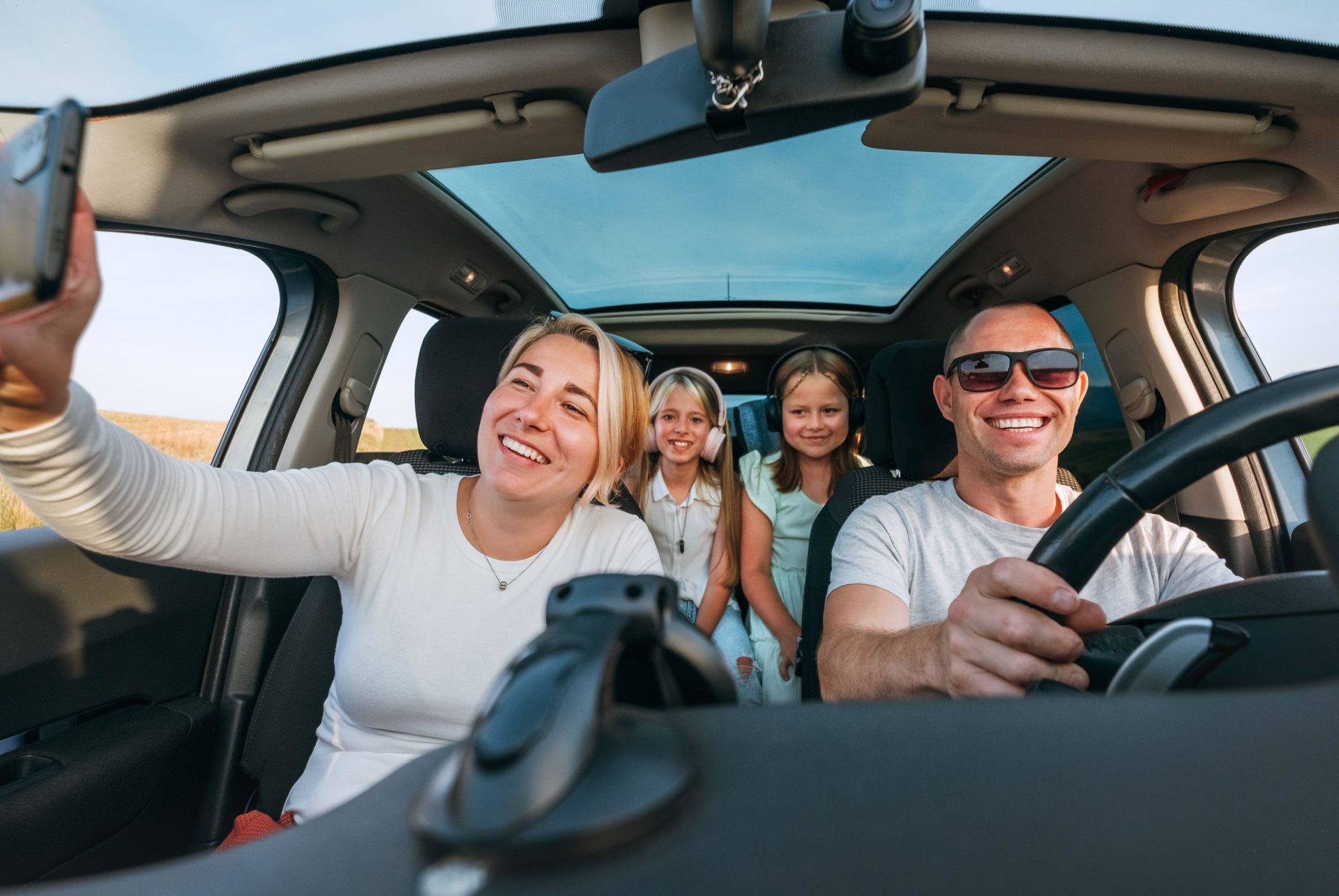 2RTD0JR Happy young couple with two daughters inside car during auto trop. They are smiling, laughing and taking selfies using smartphone. Family values, trav