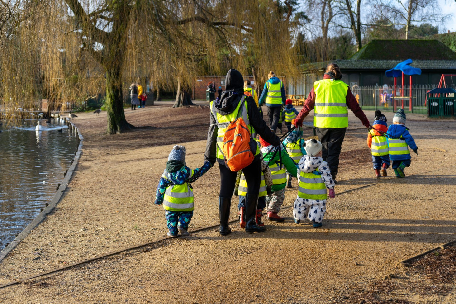 A group of children from a nursery school in high visibility jackets walking with teachers or childcare workers through a park in winter