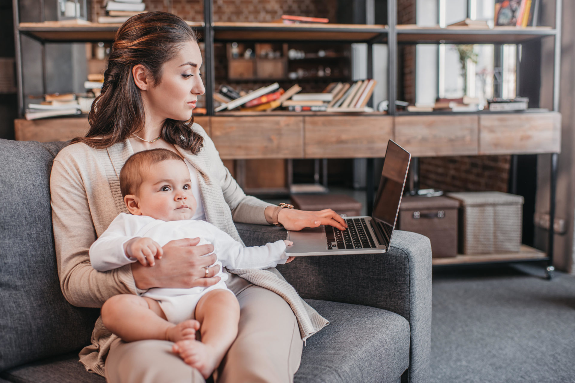 A busy woman working on her laptop while holding her baby in her lap. Image: LightField Studios Inc. / Alamy Stock Photo