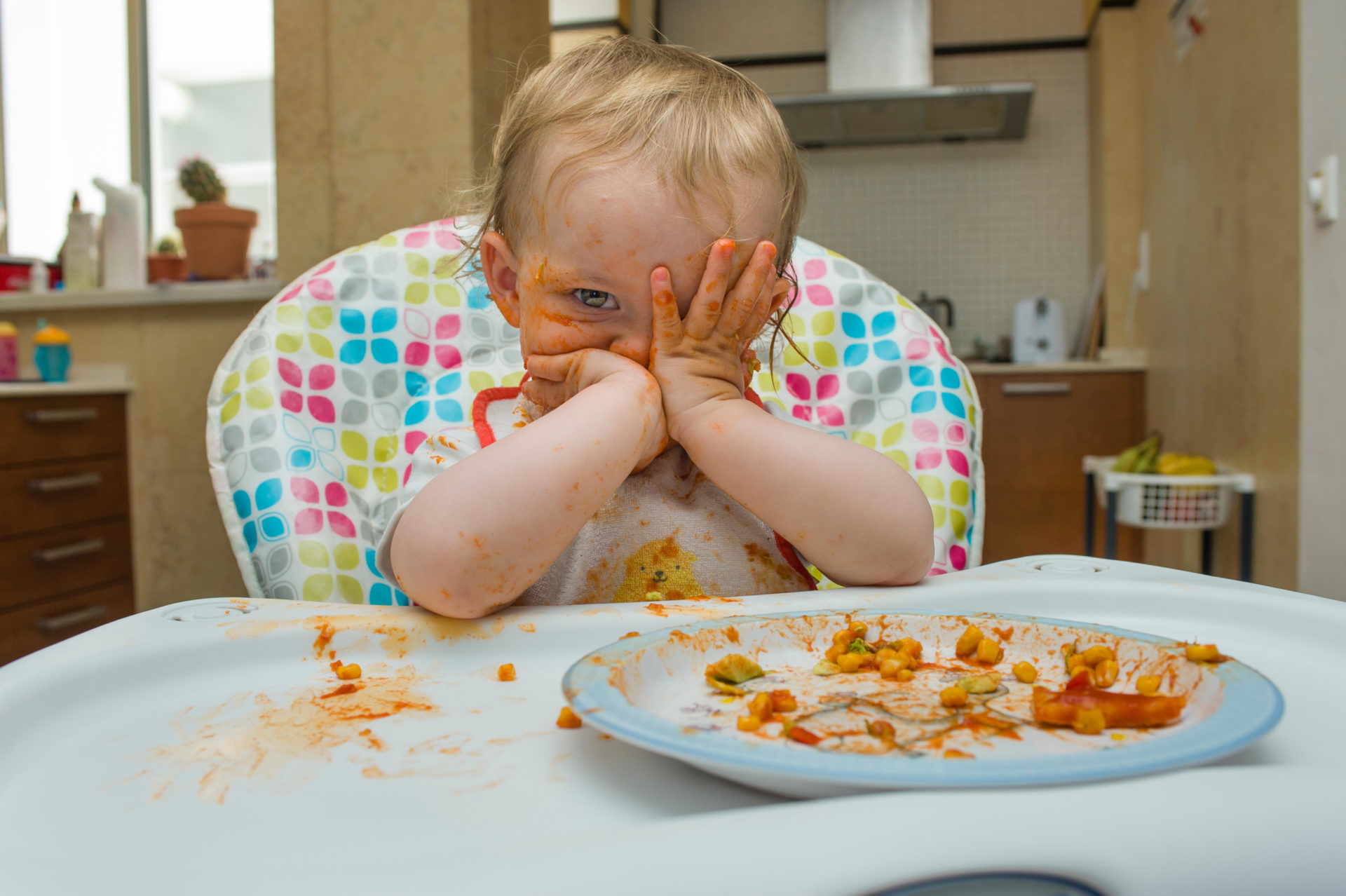 A baby girl (age 15 months) eats a meal of sweet corn and tomato sauce in a messy manner and plays with her food on a high chair