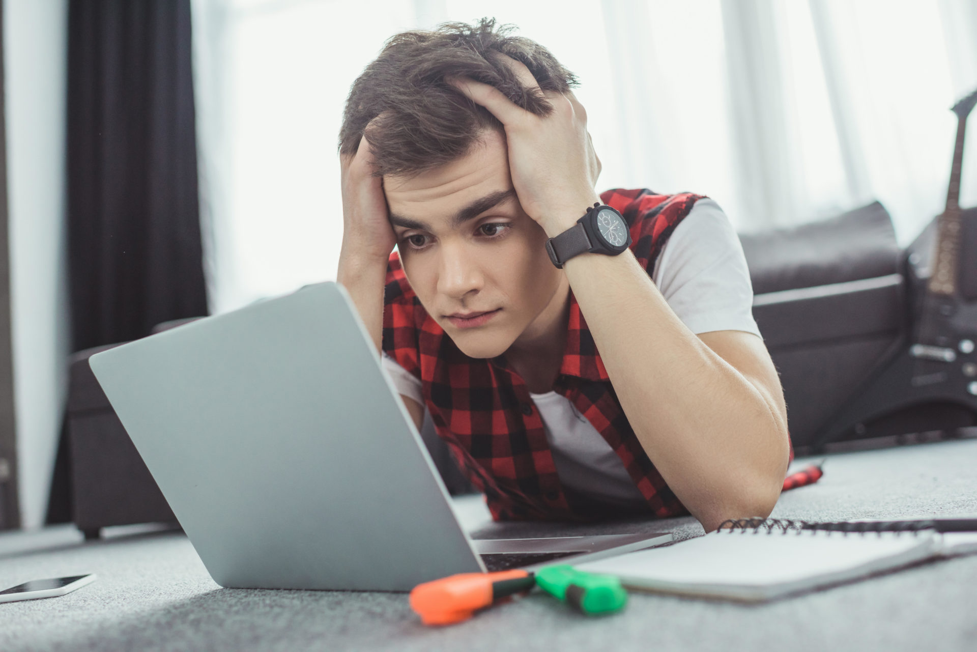 Stressed teen boy studying with laptop while lying on floor