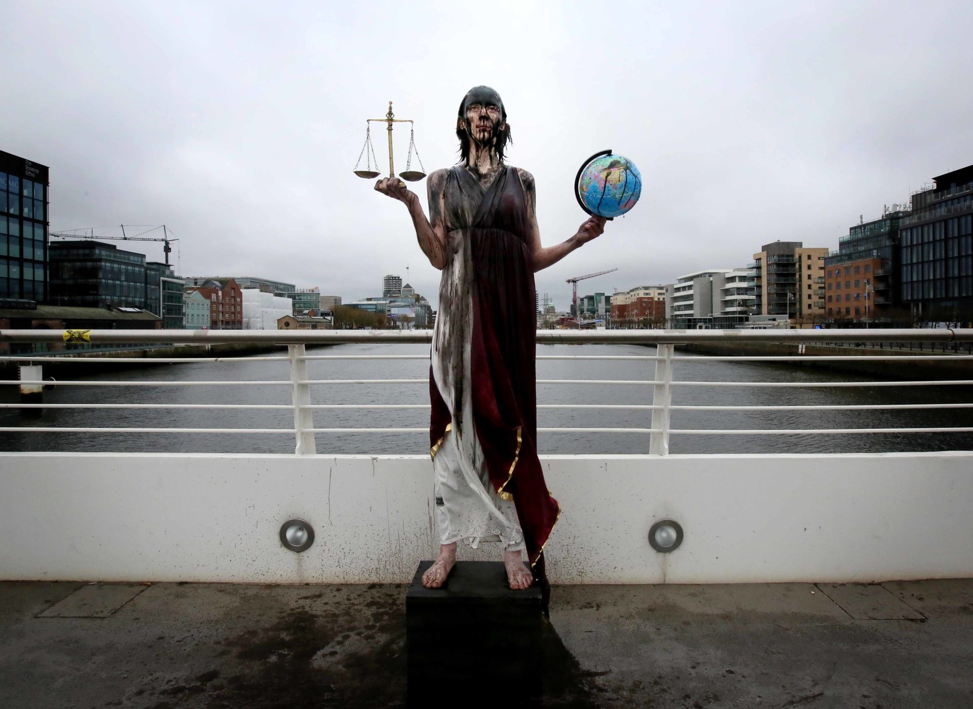 A woman poses as Lady Justice. She is covered in oil and standing on a bridge across the Liffey in Dublin City. In her left hand she holds a set of scales, in her right a globe. Both are also covered with oil.