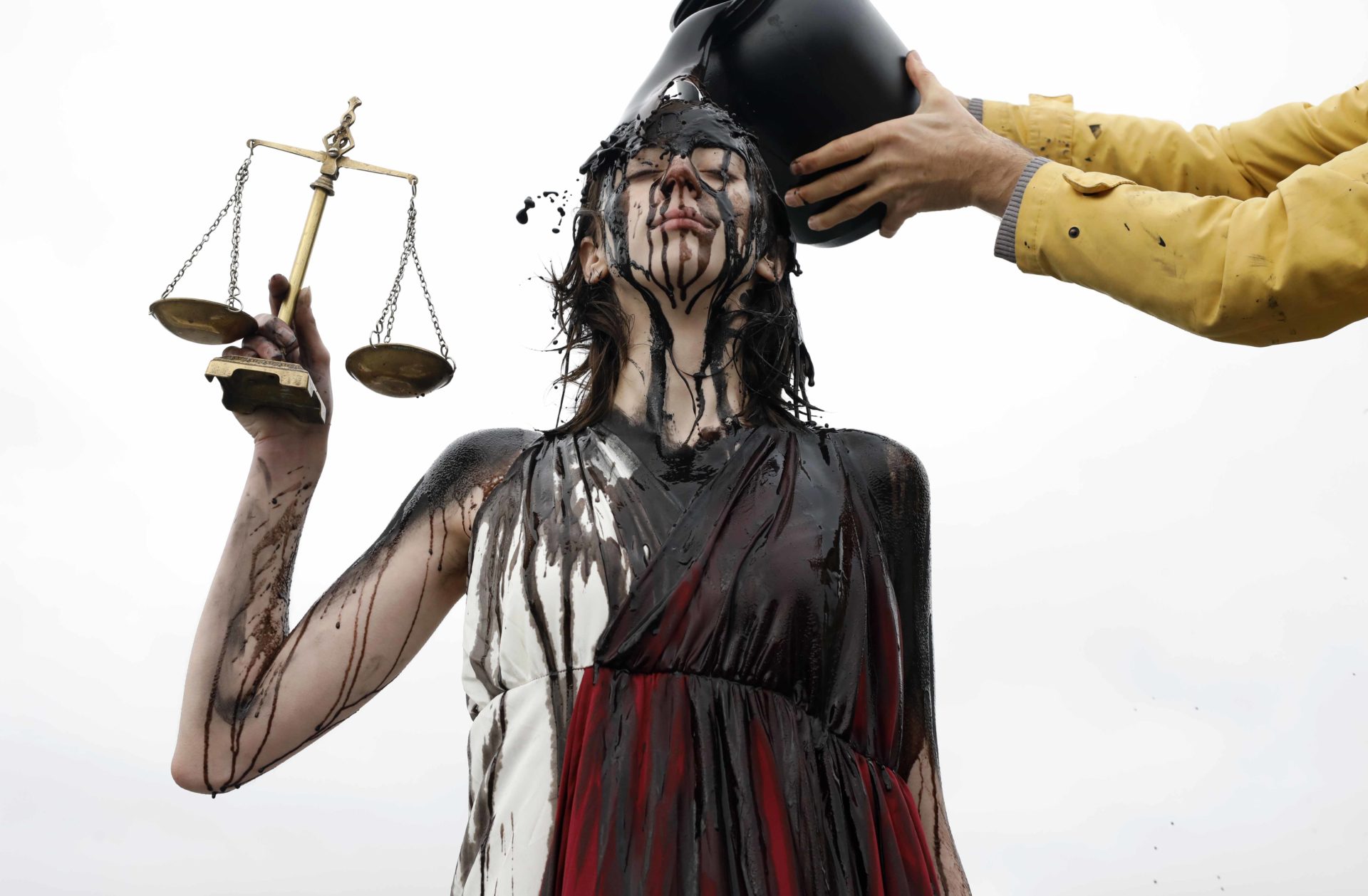 A woman poses as Lady Justice. Two hands wearing a yellow coat can be seen pouring oil over her head. In her left hand she holds a set of scales.