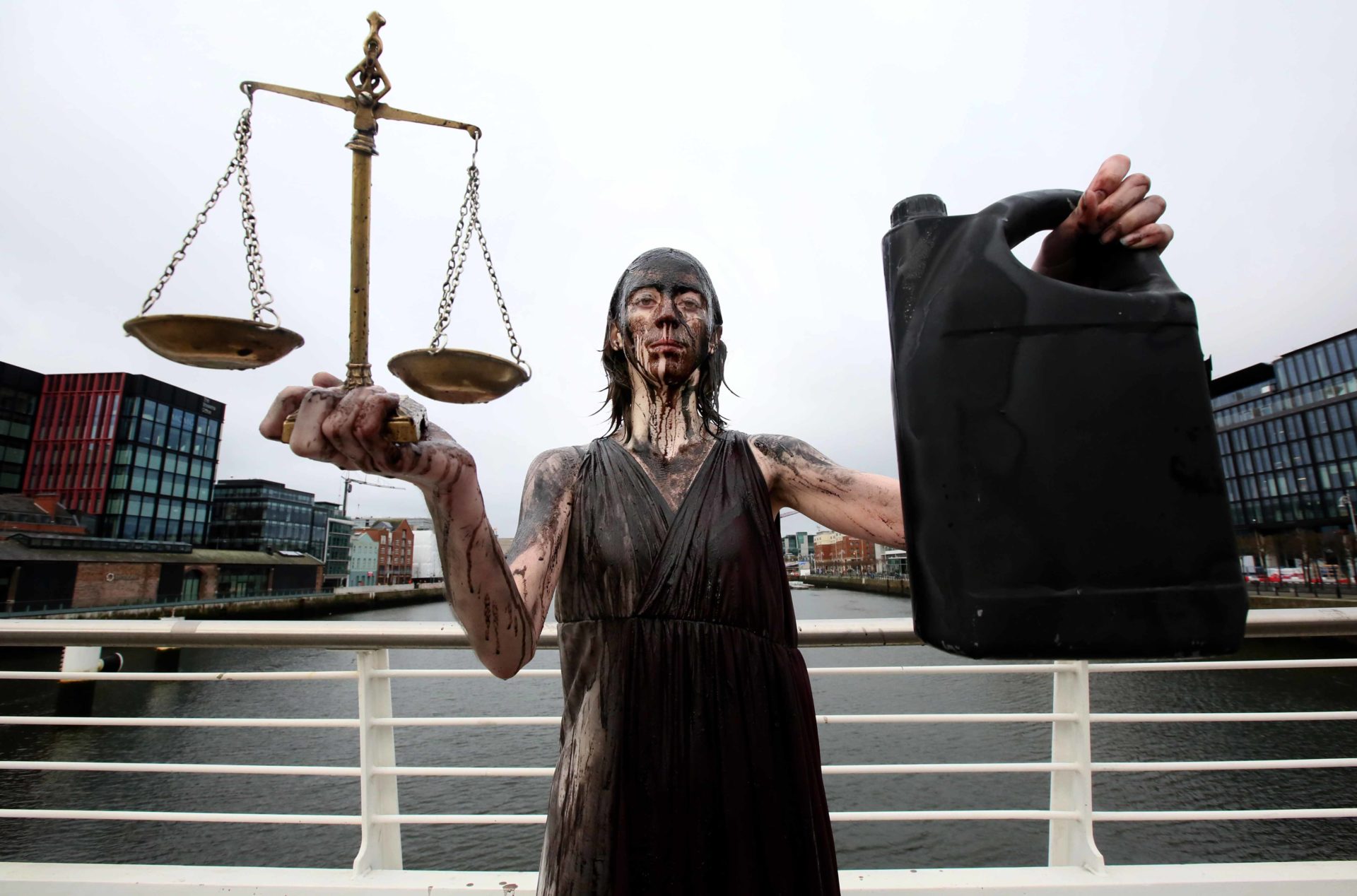 A woman stands, dressed as Lady Justice and covered in oil on a bridge crossing the Liffey in Dublin city. In her left hand she holds out a set of scales to the camera, in her right she holds a jug of oil.