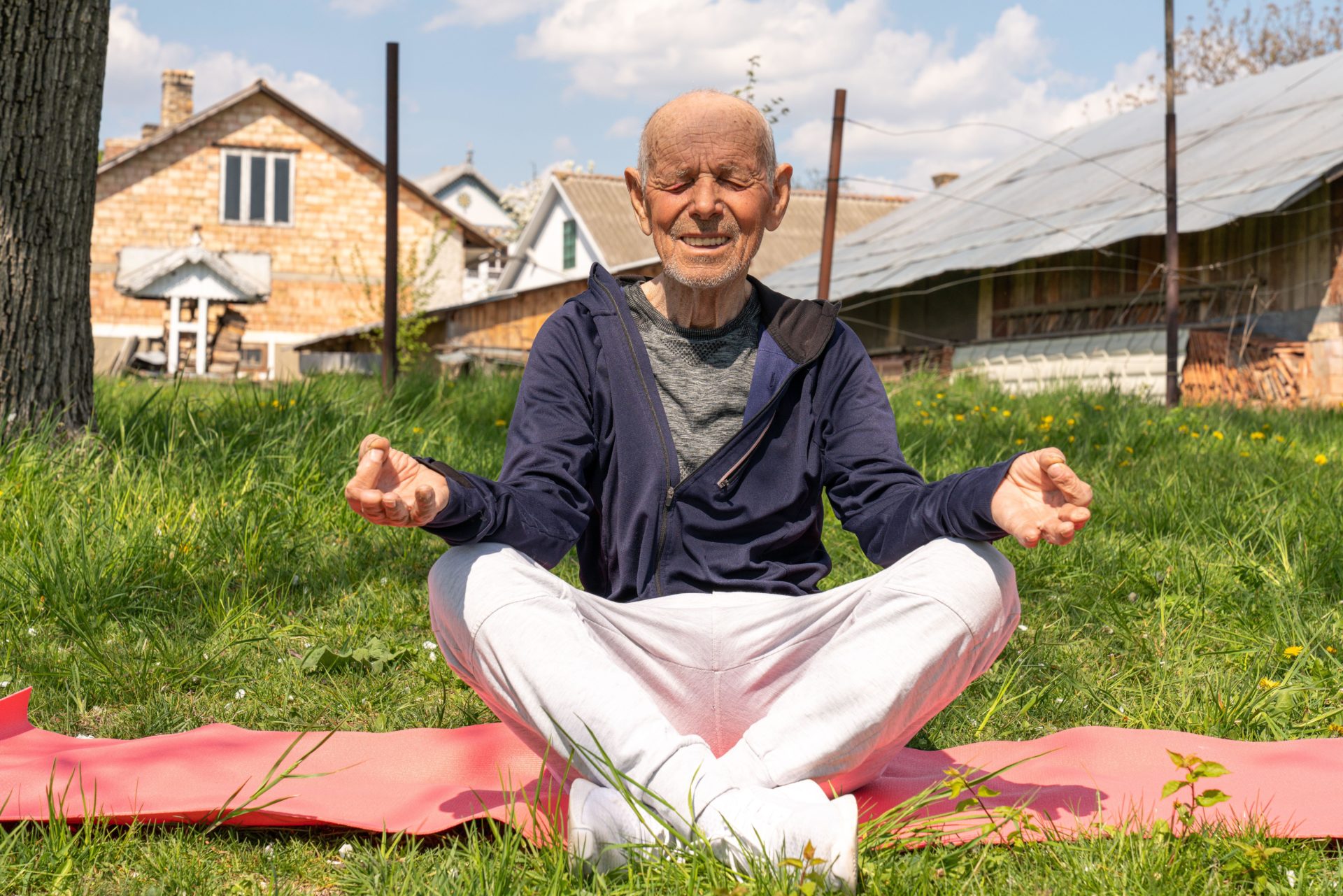 Elderly man sitting in lotus position on yoga mat in the backyard.