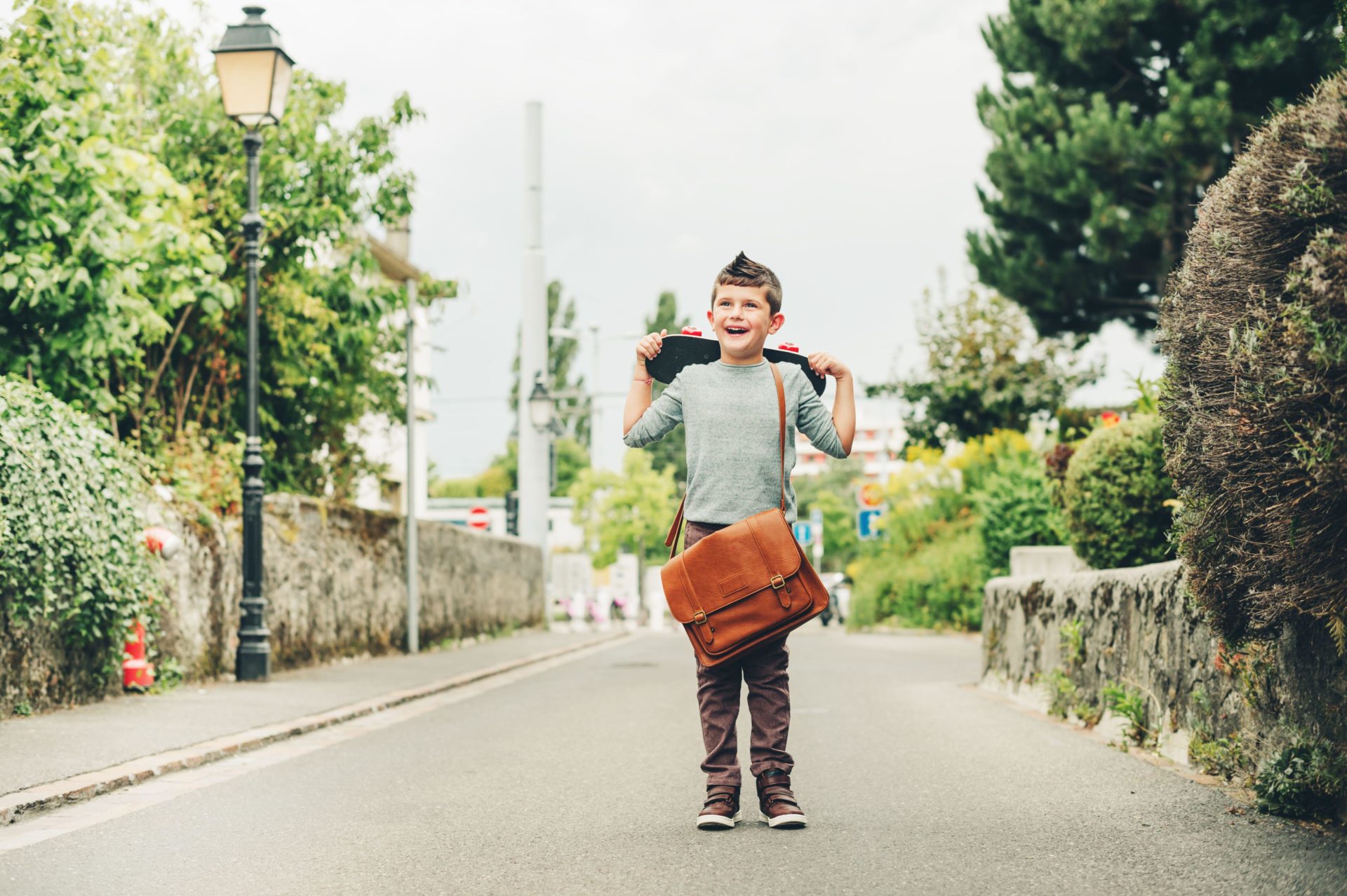 Outdoor portrait of schoolboy wearing brown leather bag over shoulder, holding skateboard. 