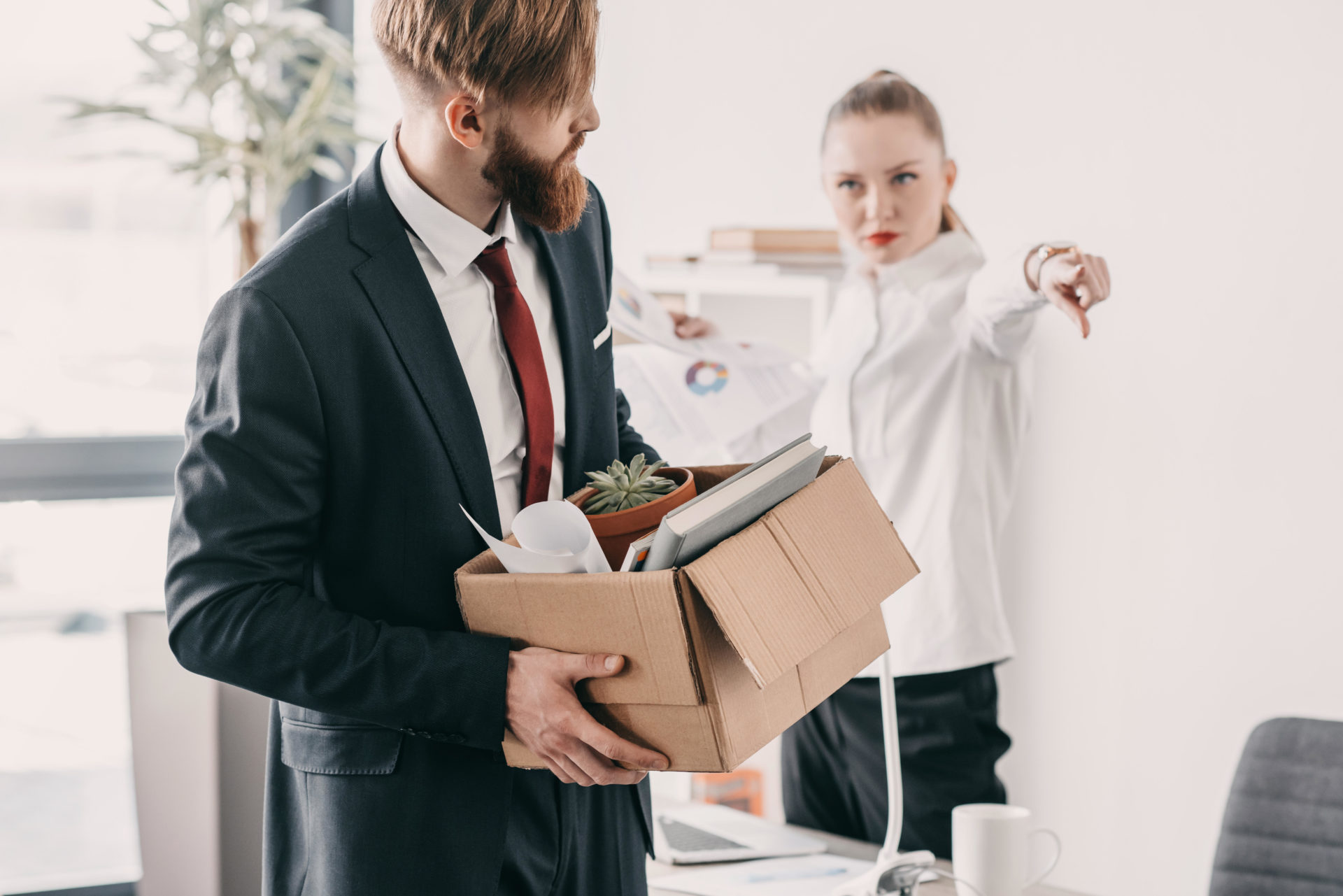 Young upset fired businessman with cardboard box and boss in office.