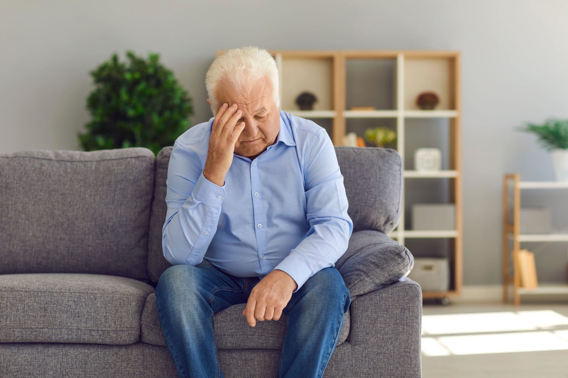 Depressed senior man sitting on sofa at home suffering from loneliness or bad headache. 