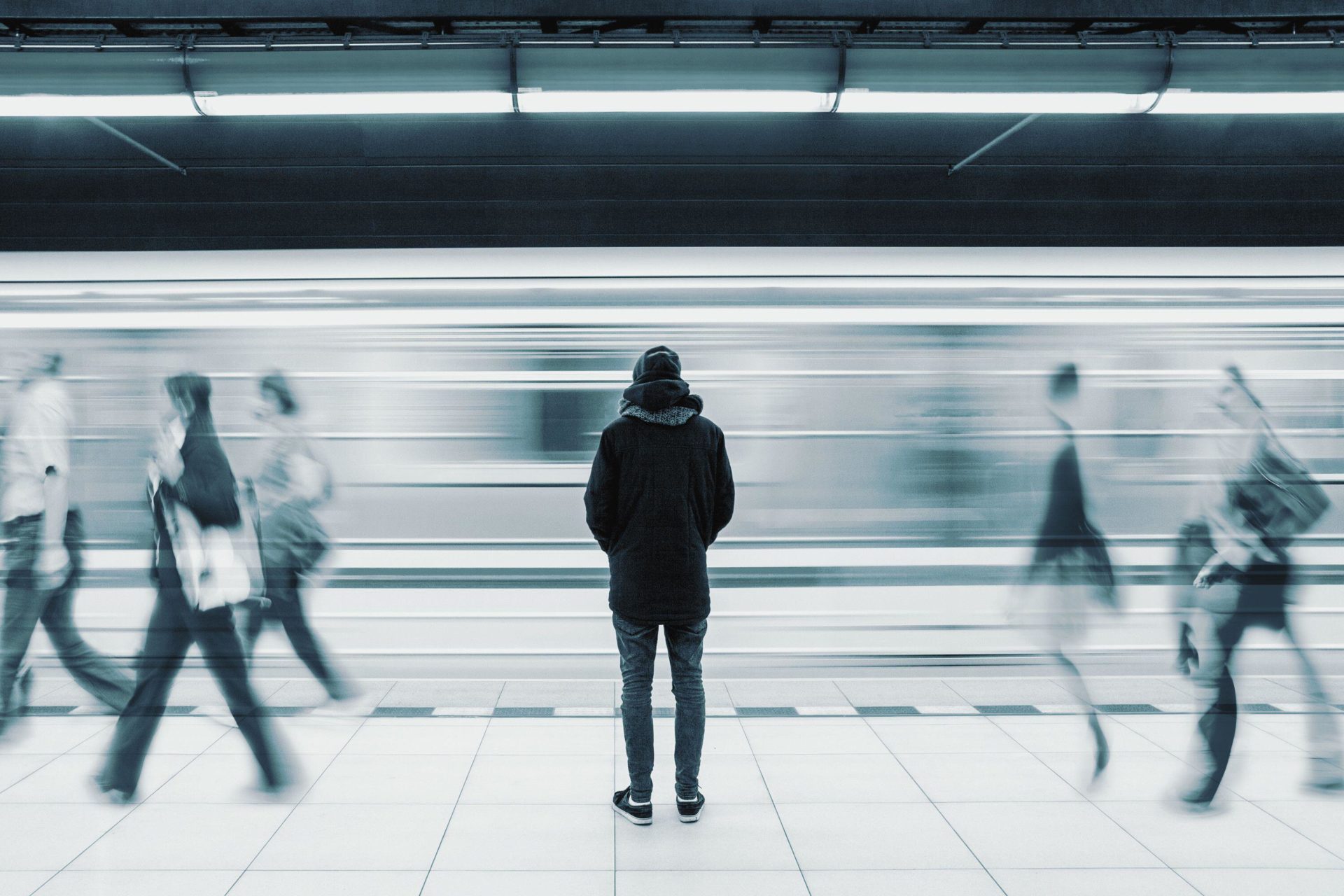 Lonely young man shot from behind at subway station with blurry moving train and walking people in background.