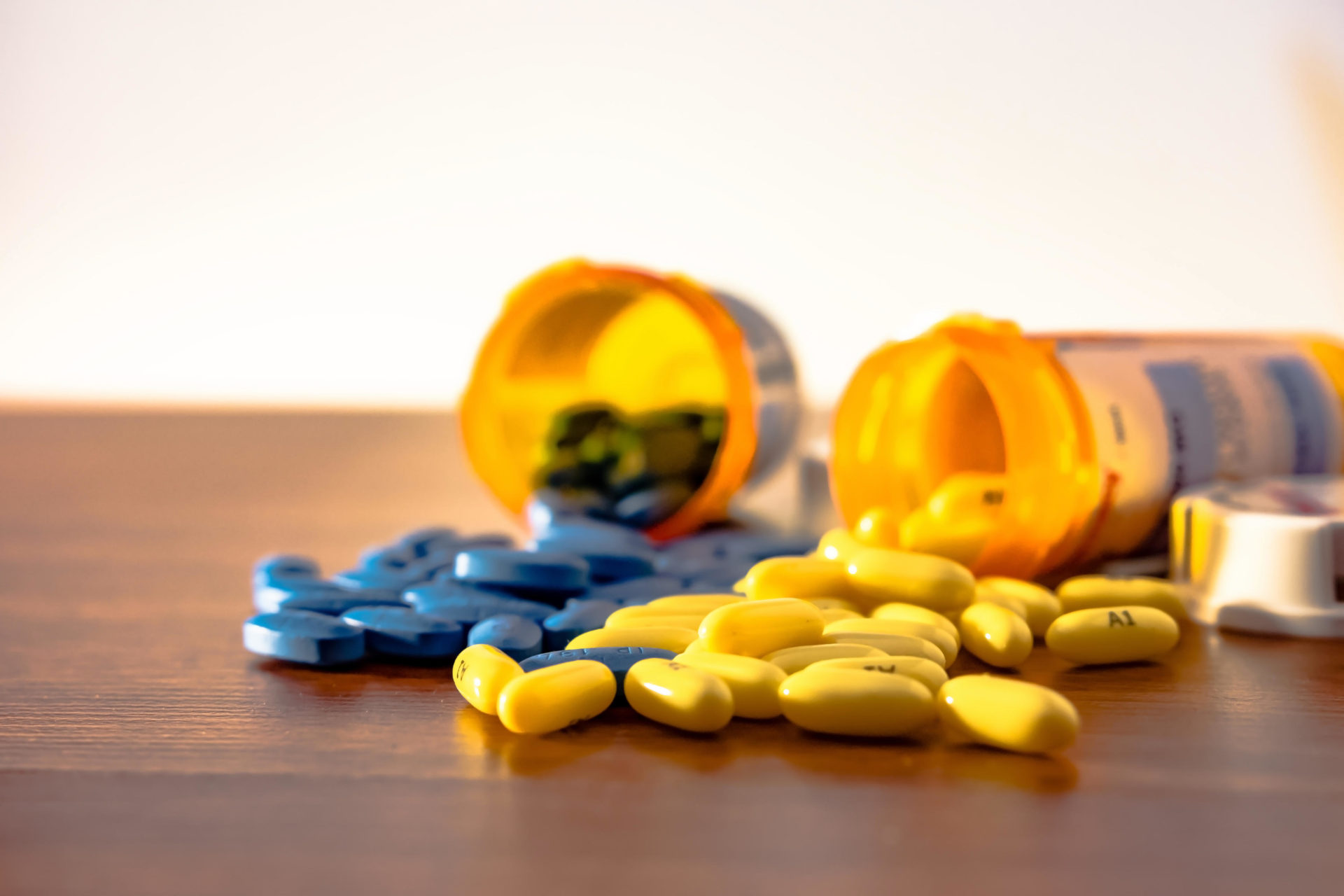 Two prescription bottles overturned with blue and yellow pills spilling onto the wooden table with white background. Image: Alamy