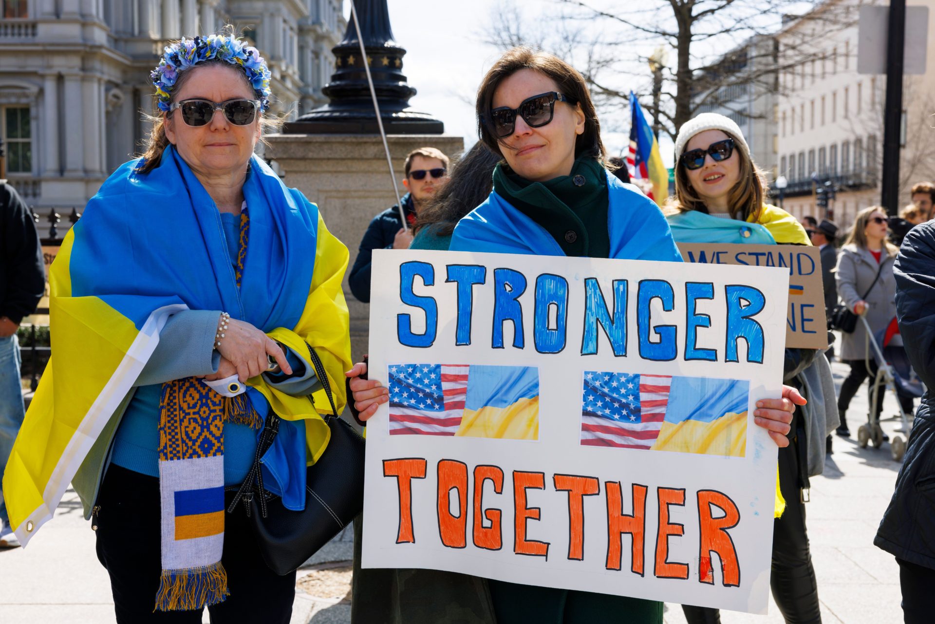 A small crowd of people hold Ukrainian flags and signs outside the White House as Ukrainian President Volodymyr Zelenskyy meets with President Trump to sign a deal granting the US access to Ukraine's rare minerals.