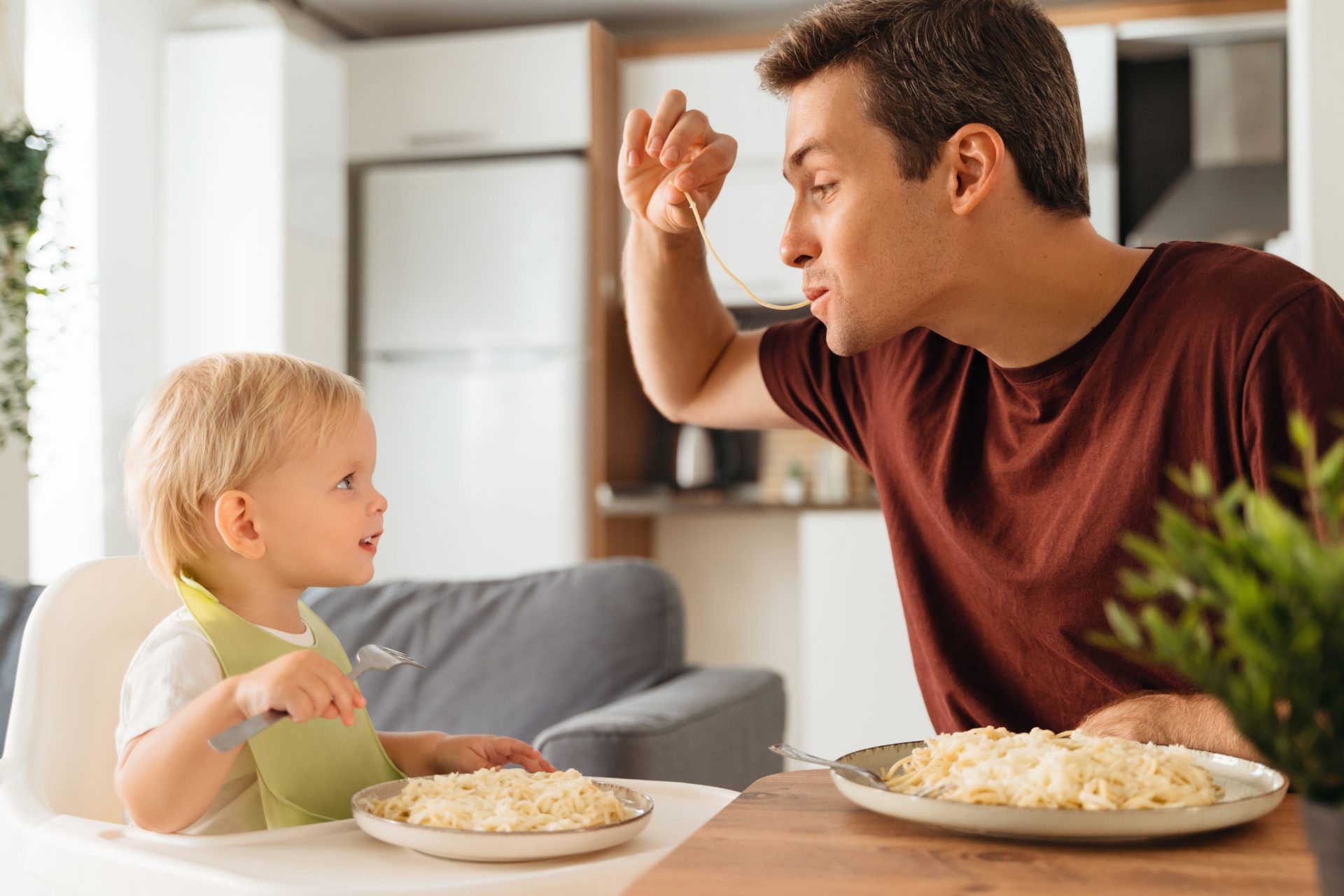 Happy dad playing with spaghetti while having dinner with his baby boy in bib learning to eat with fork looking at funny dad with interest. 