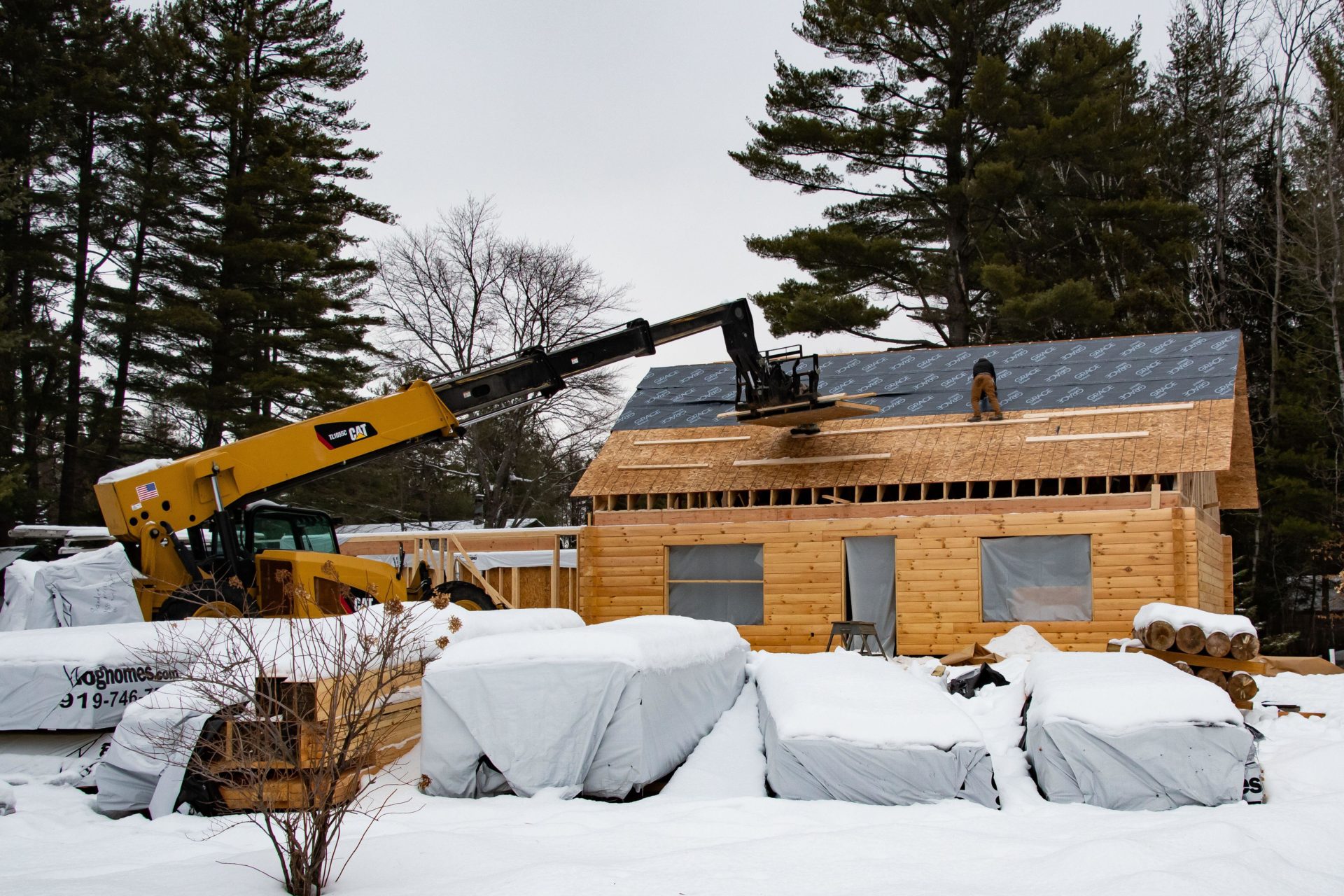 Two construction workers installing a roof covering on a manufactured log home in winter with a Cat TL1055C Telehandler in Speculator, NY USA .