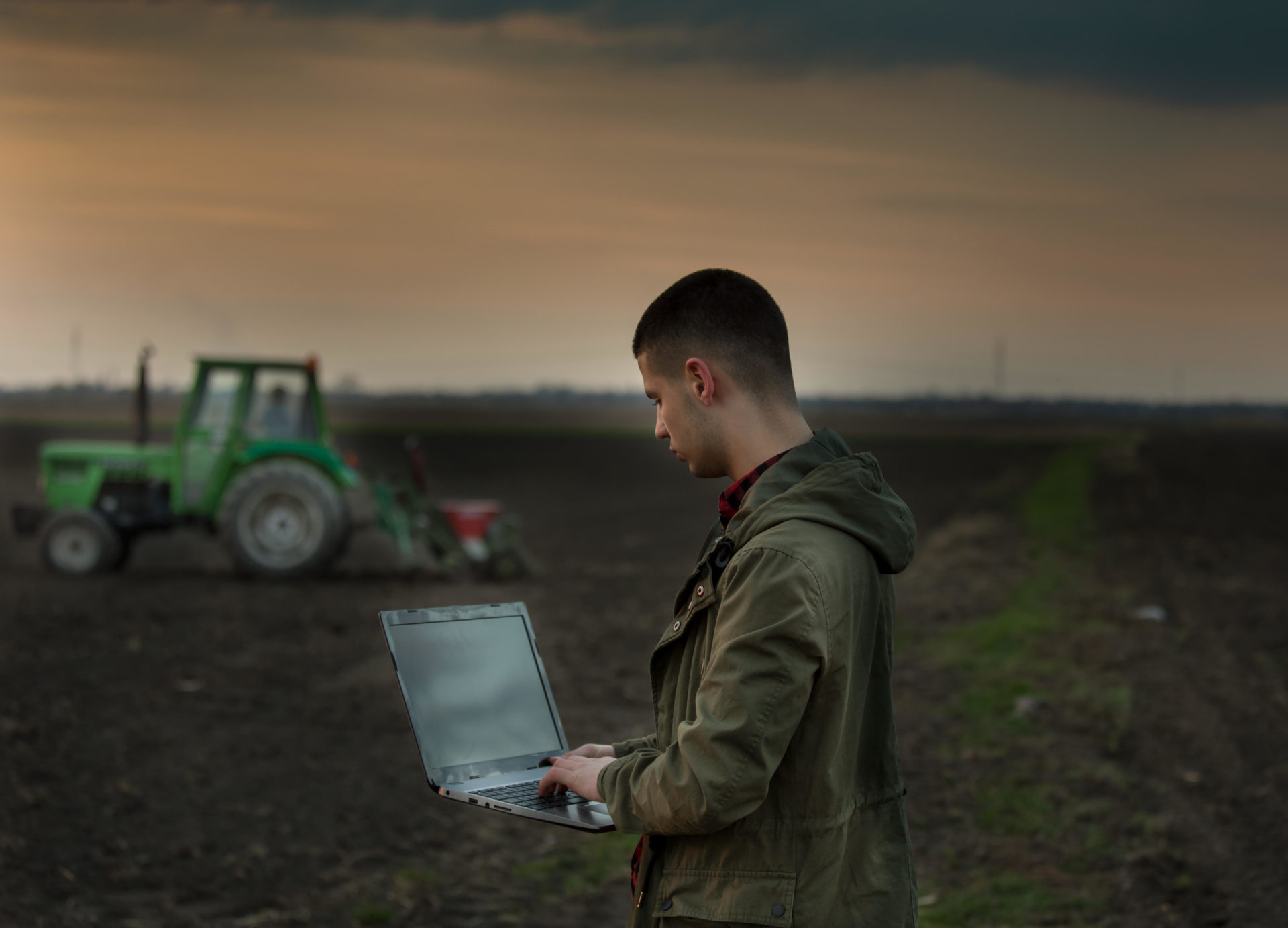 Young farmer with laptop standing in field with tractor harrowing in background.