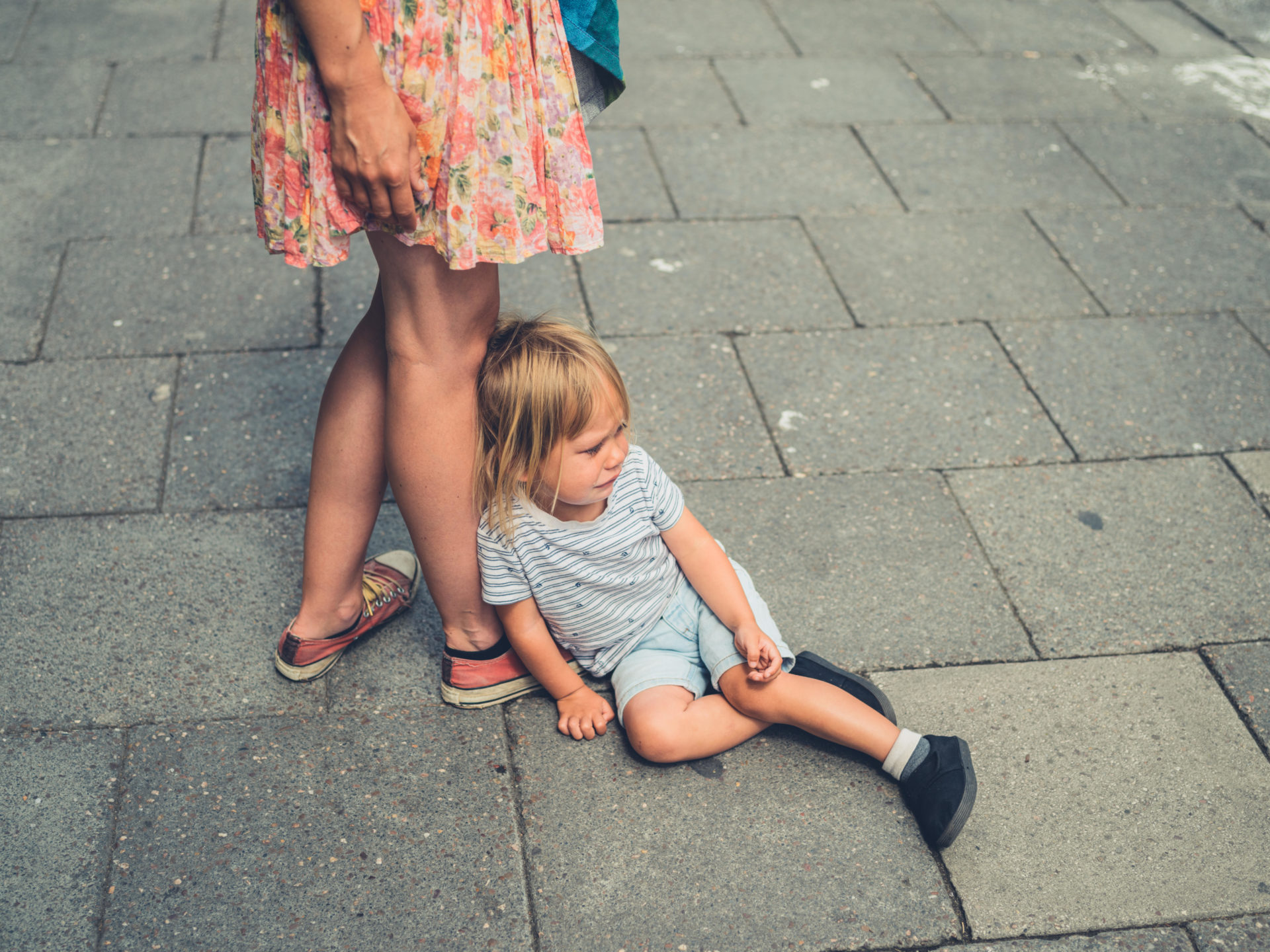 A sad crying toddler is sitting in the street by his mother's feet. 