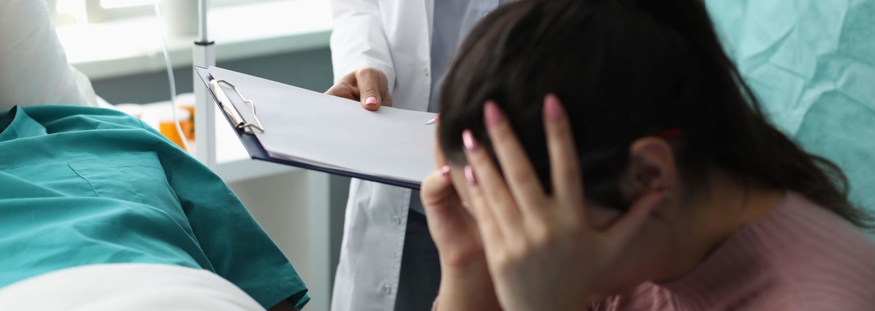 Doctor holding documents in front of crying wife and patient in hospital room.