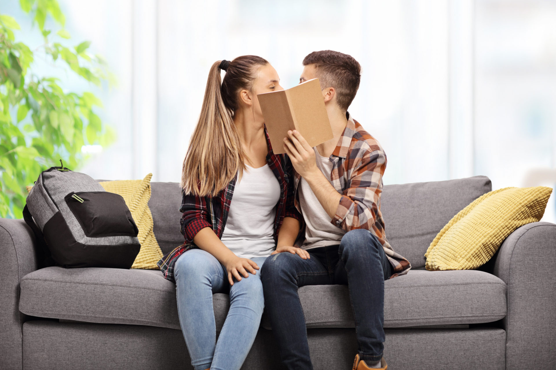 Teenage couple kissing on a sofa and hiding behind a book.