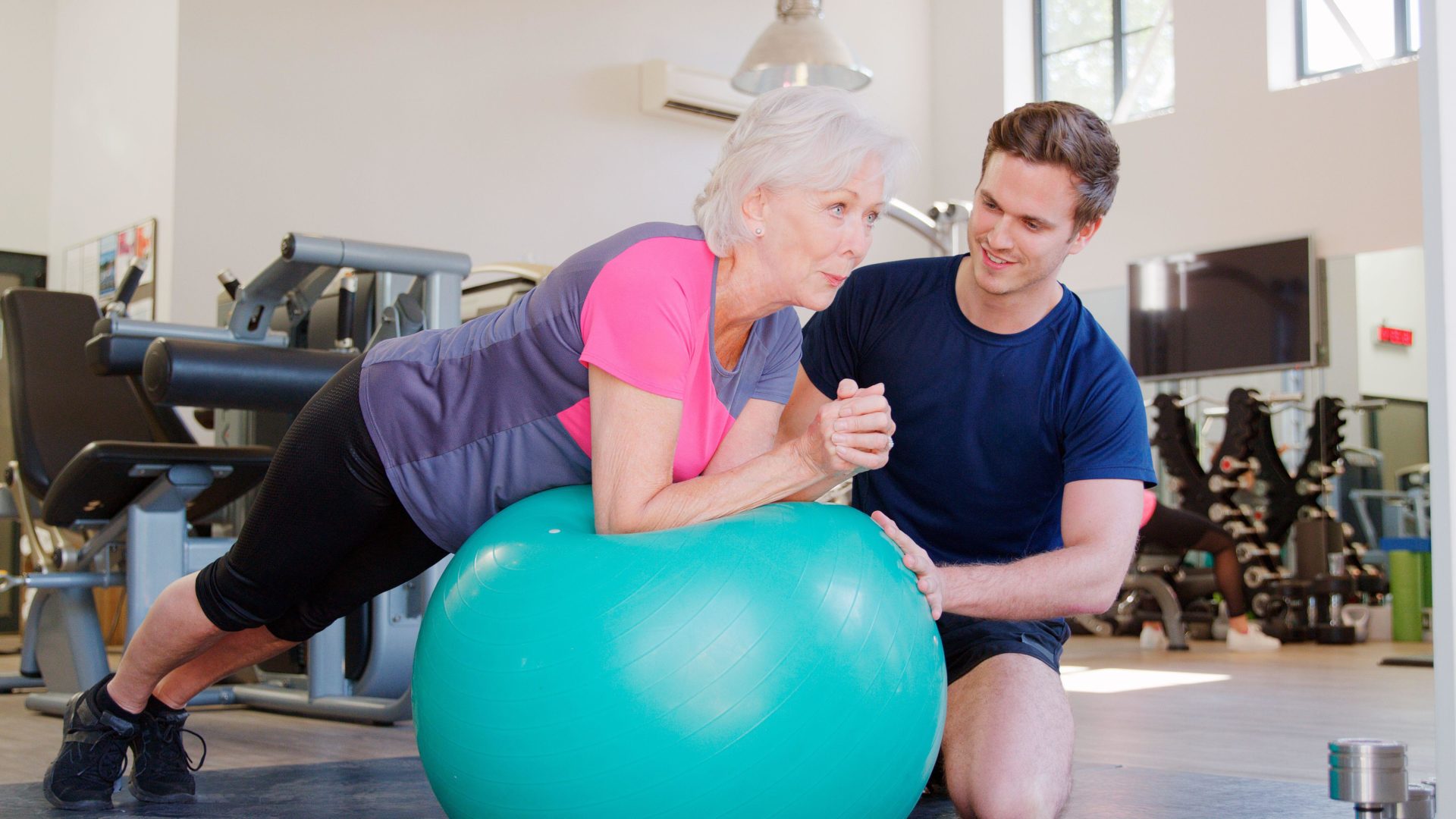 3BB0CHY Senior Woman Exercising On Swiss Ball Being Encouraged By Personal Trainer In Gym