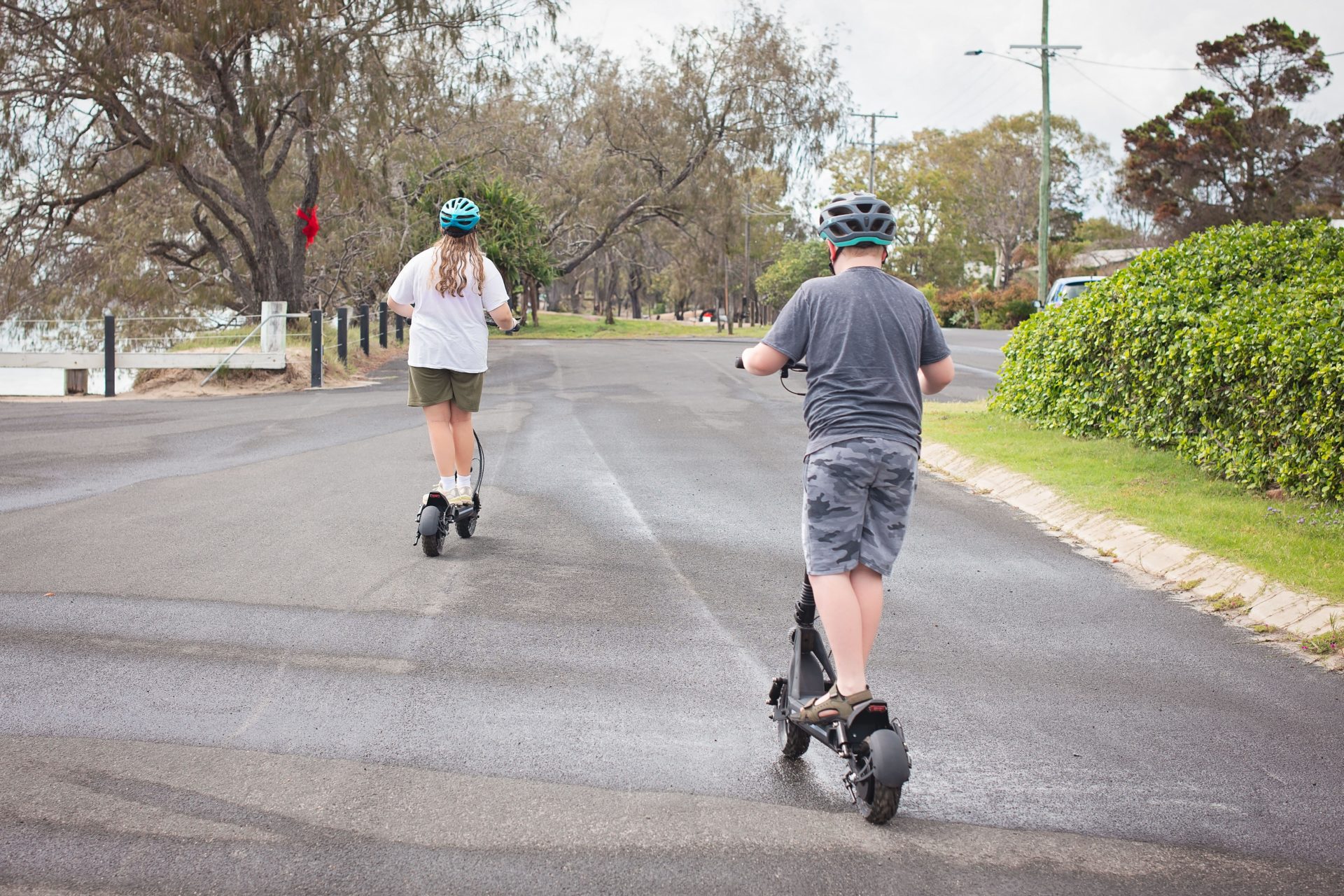 2YY8JT6 pre teen kids on e-scooters wearing helmets paved road riding away
